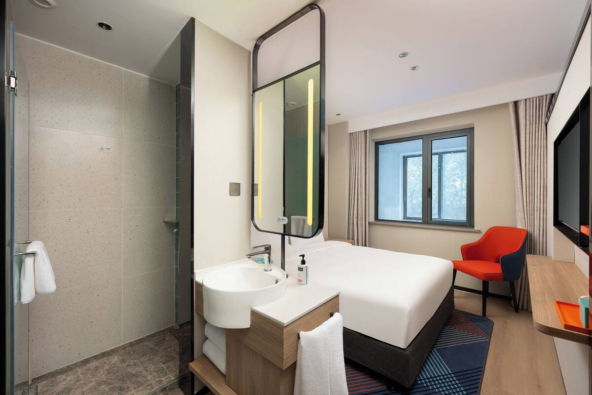 A hotel room with a large bed, a bathroom with a sink and mirror, a shower, a window with curtains, and a desk with a chair.