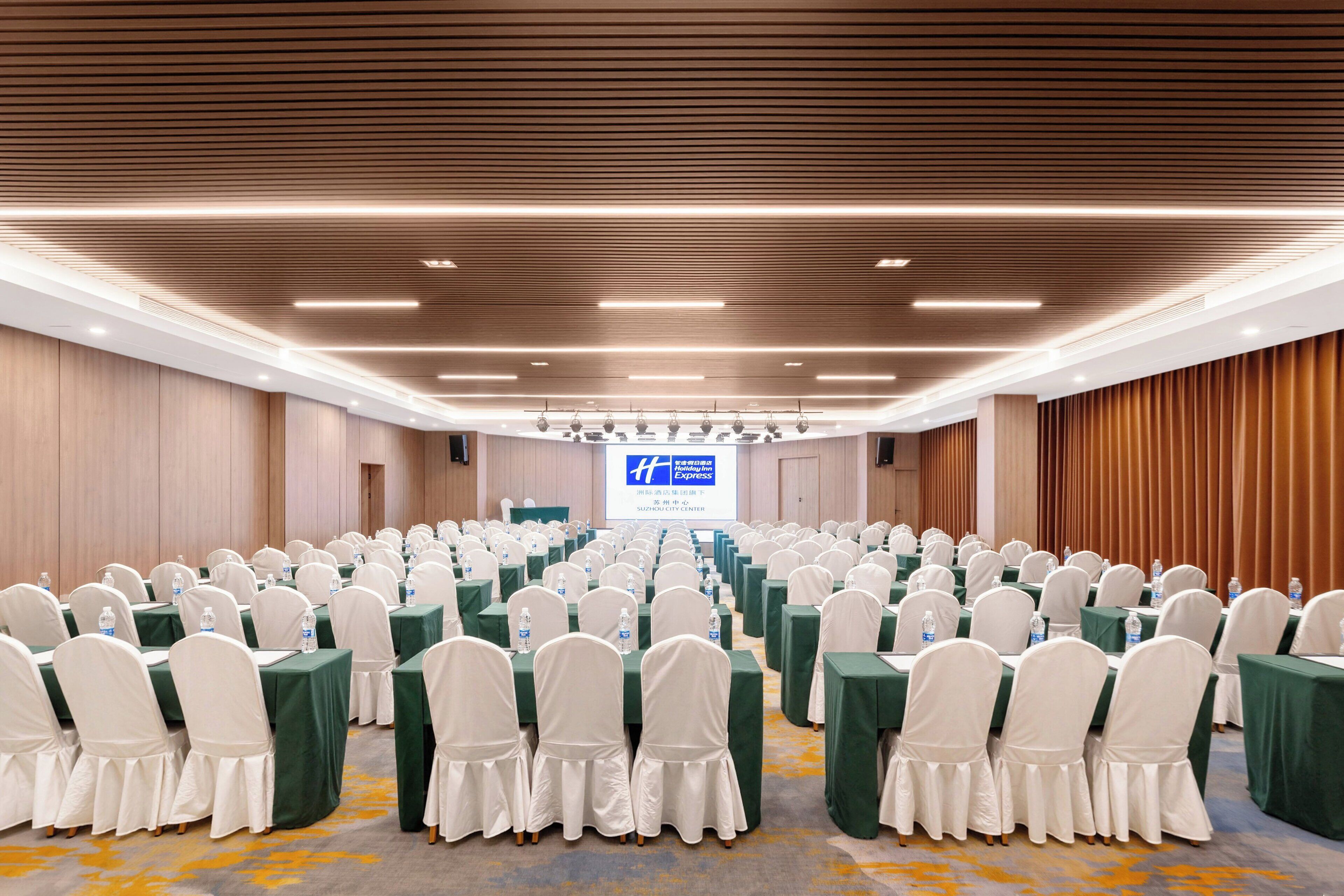 A conference room with rows of tables and chairs, a podium, and a screen displaying text.