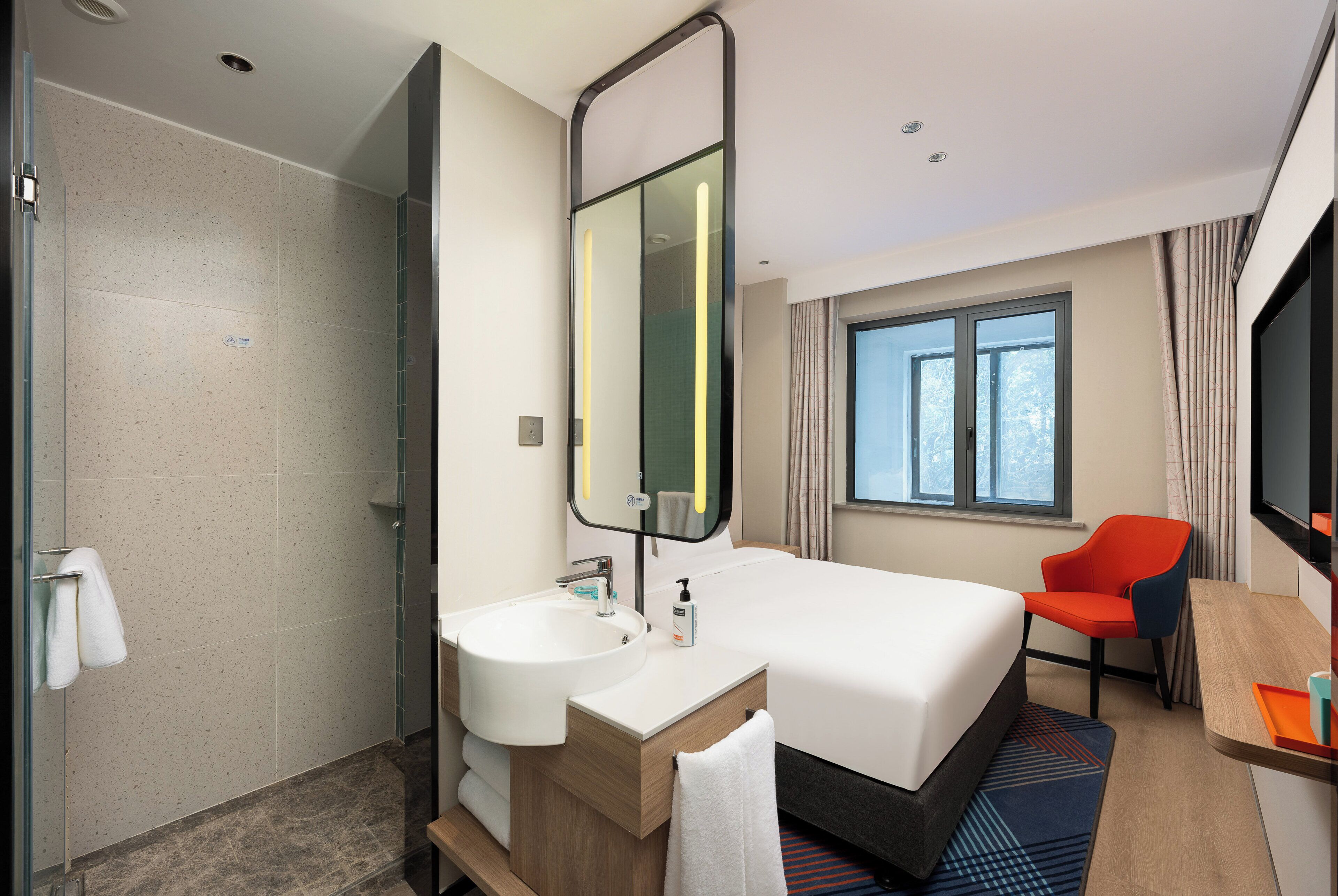 Una habitación de hotel con una cama grande, un baño con lavamanos y espejo, ducha, una ventana con cortinas y un escritorio con silla.