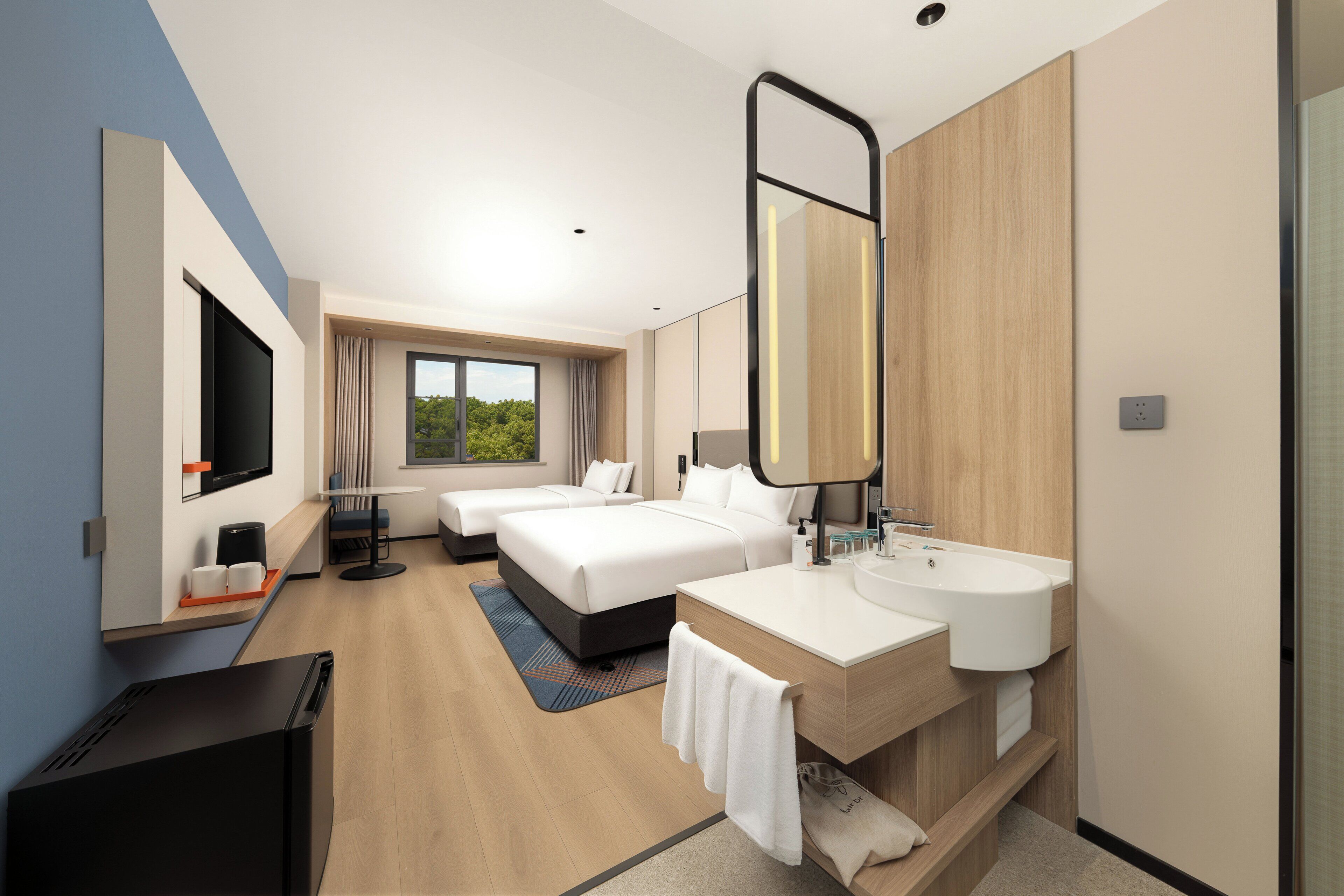 A hotel room with a large bed, two bedside tables, a TV, a bathroom with a sink and mirror, and a window with a view of trees.