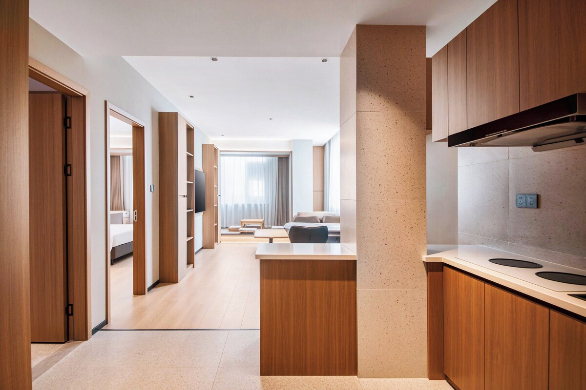 Een moderne hotelkamer met een houten vloer, een groot raam met gordijnen, een flatscreen televisie en een kitchenette met fornuis en magnetron.