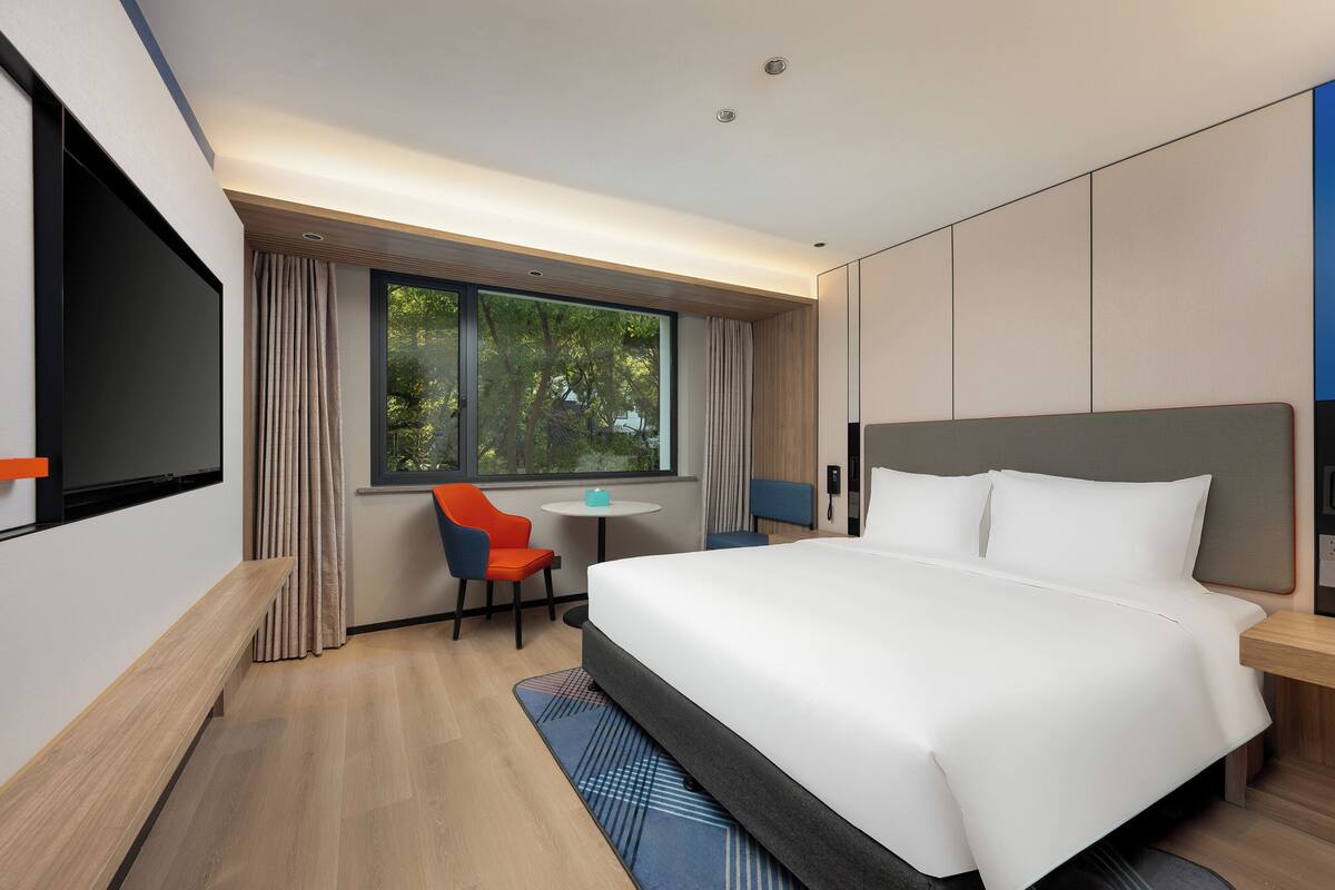 A modern hotel room with a large bed, a desk, a chair, a TV, and a window with curtains.