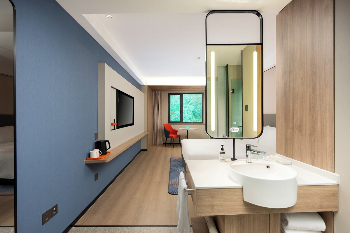 Een moderne hotelkamer met een grote spiegel, twee witte wastafels, een houten opbergmeubel, een televisie en een rode stoel.