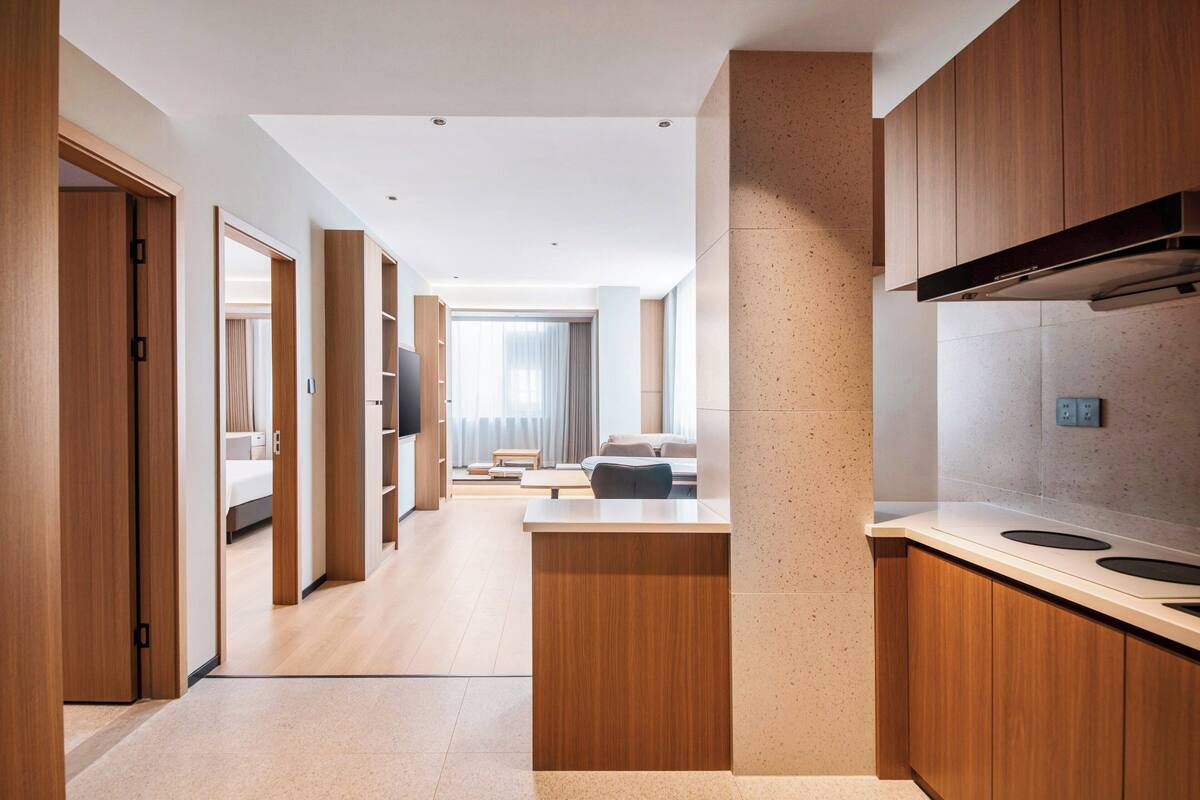 A modern hotel room with a wooden floor, a large window with curtains, a flat-screen TV, and a kitchenette with a stove and microwave.