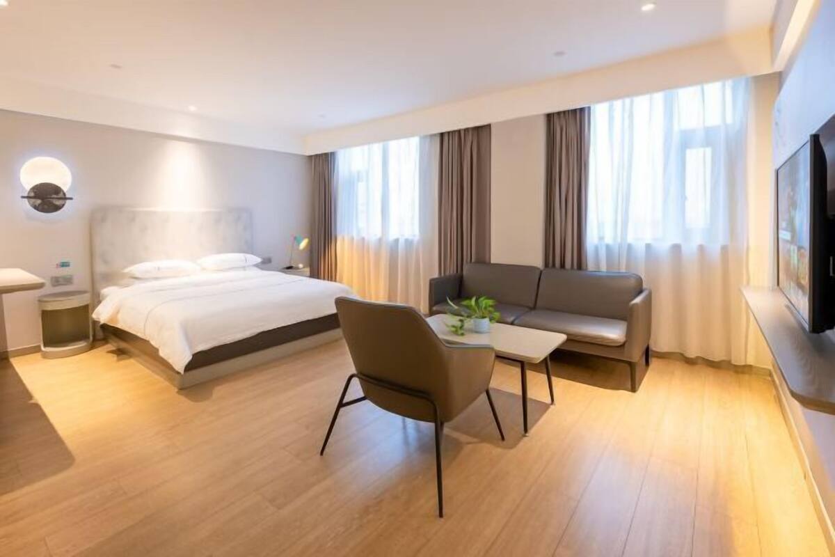 A modern hotel room with a large bed, a sofa, a small table with a plant, and a TV mounted on the wall.