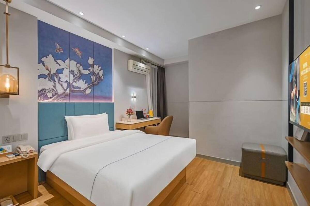 A modern hotel room with a bed, a desk, a chair, a TV, and a painting on the wall.