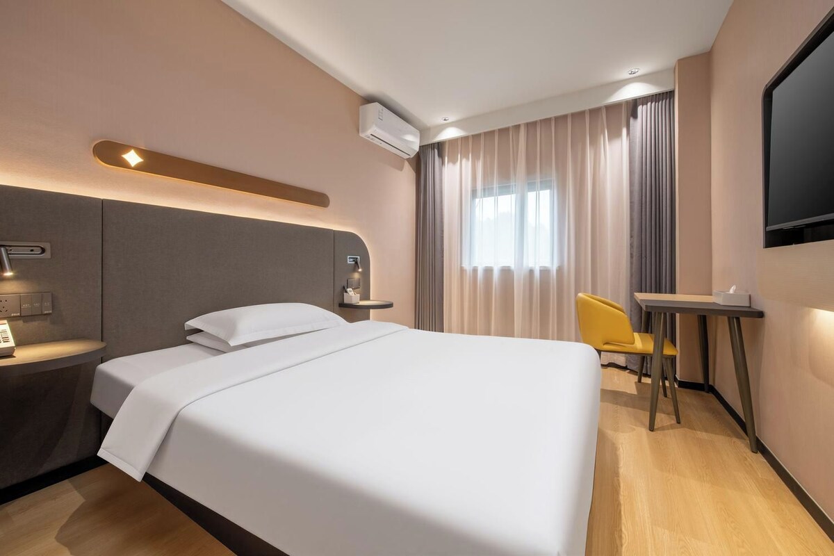 A hotel room with a bed, bedside table, lamp, air conditioning unit, window with curtains, and a desk with a chair.
