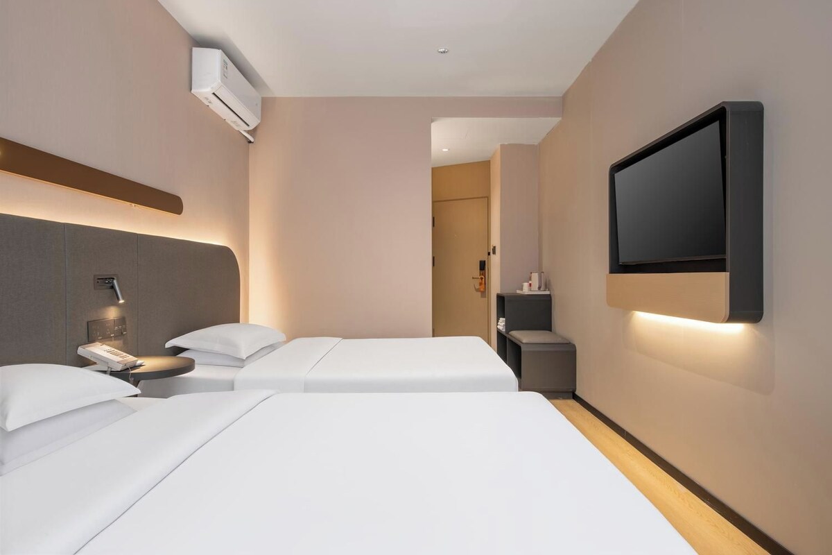 A hotel room with two beds, a TV mounted on the wall, a bedside table with a lamp, and an air conditioning unit.