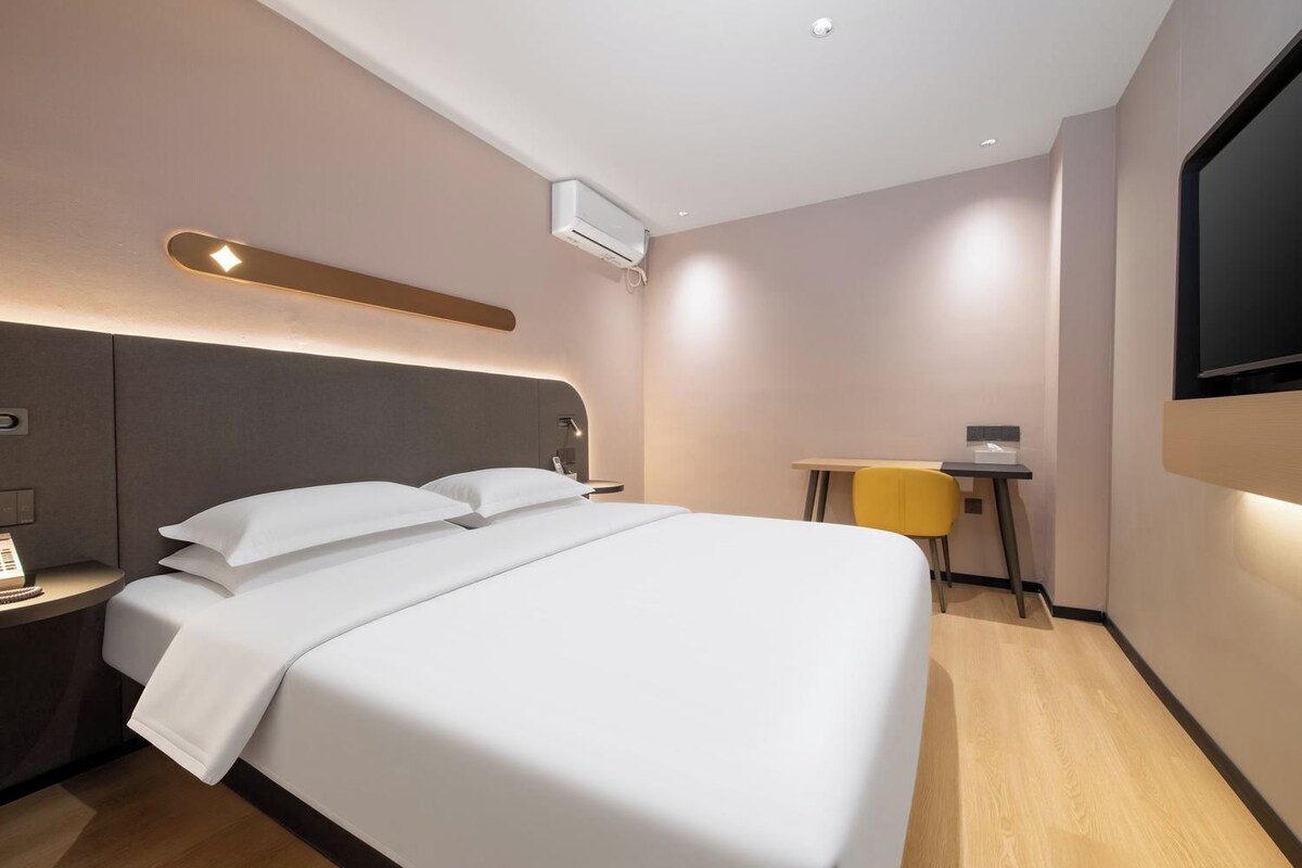 A modern hotel room with a large bed, bedside table, and a desk with a chair.