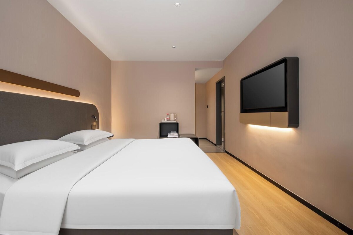 A modern hotel room with a large bed, a flat-screen TV mounted on the wall, and a bedside table with a lamp.