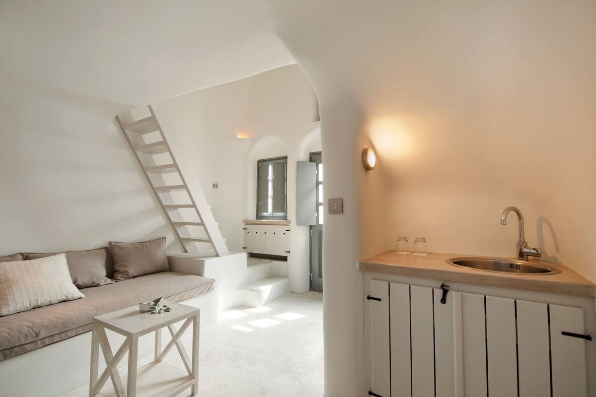 A modern, minimalist interior with a sofa, a small table, a kitchen area with a sink, and a staircase leading to another room.