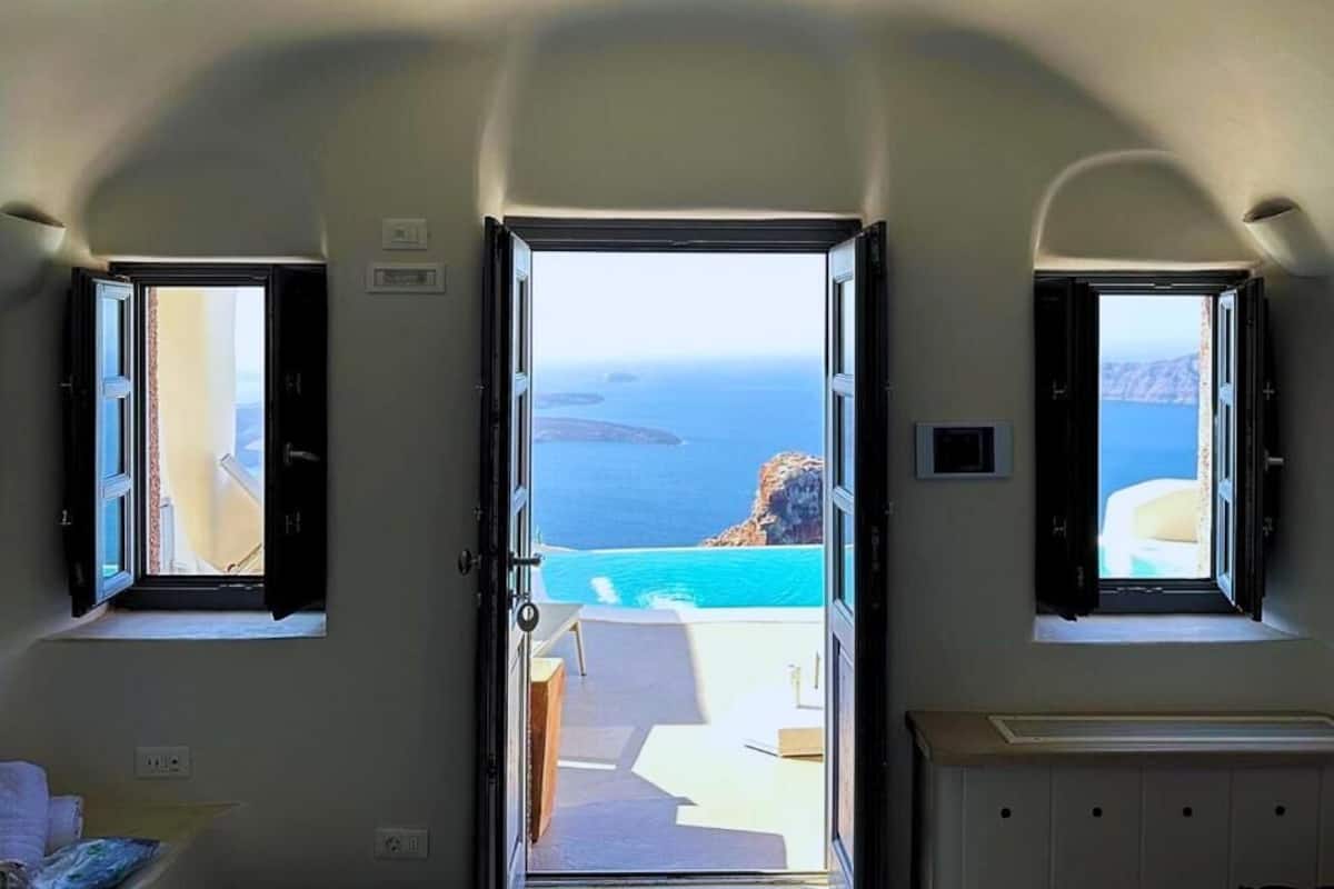A room with two open windows and a door leading to a balcony with a view of the sea.