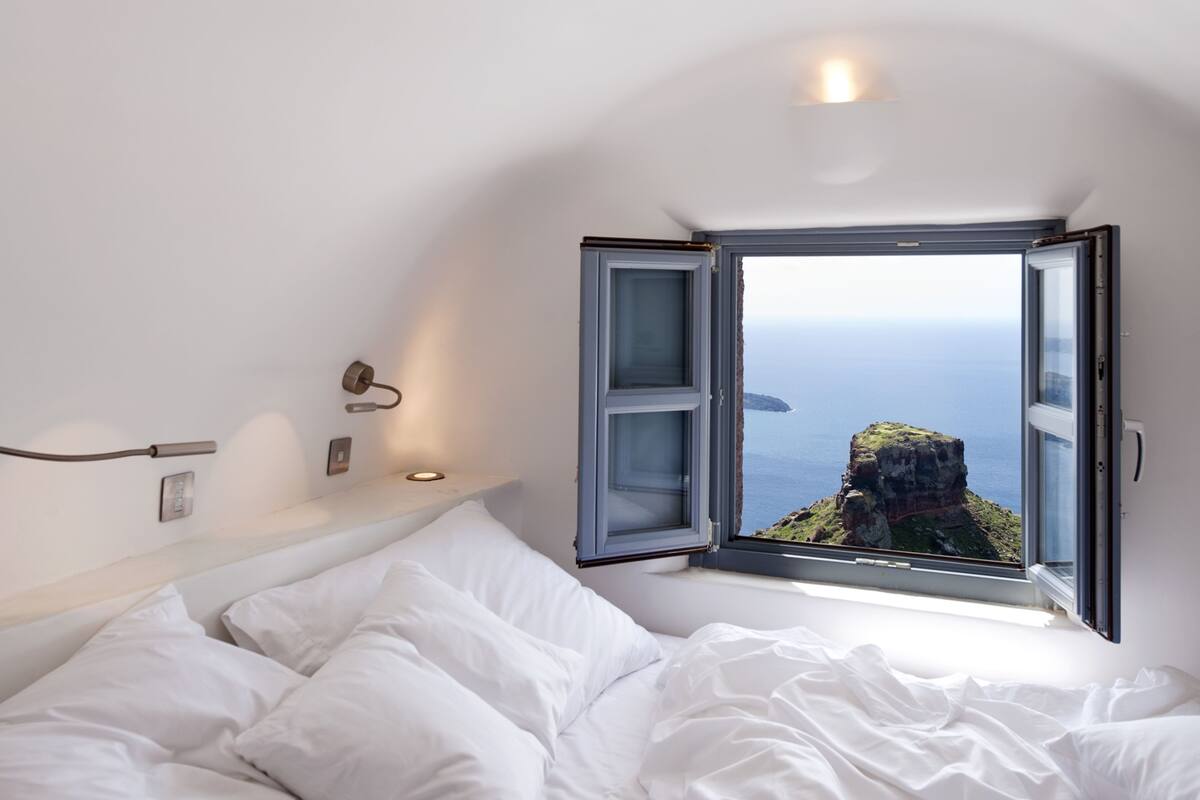 A bedroom with a large window offering a view of a cliff and ocean.