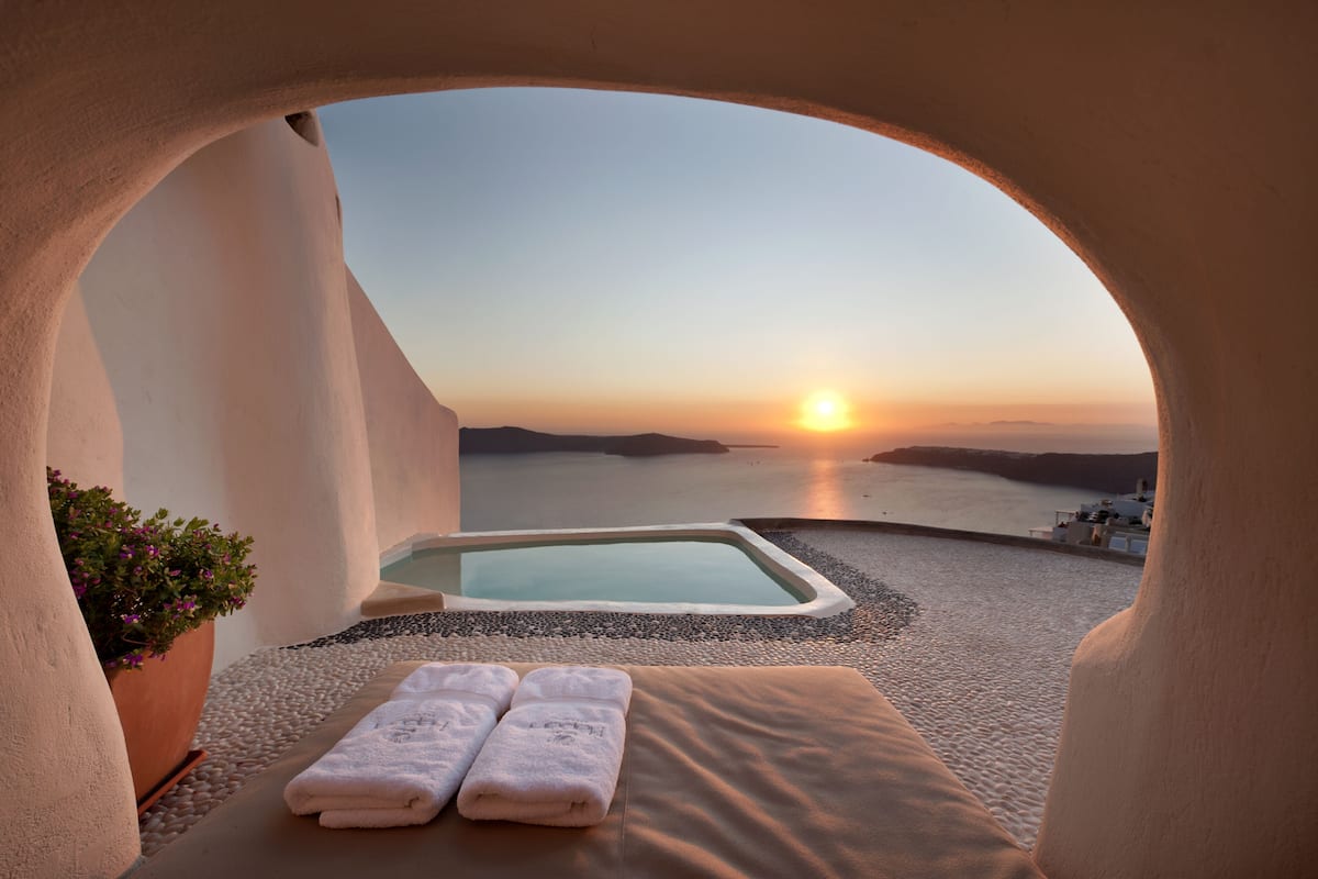 A sunlit room with a view of the sunset over water, a small pool, and a lounge area with towels.