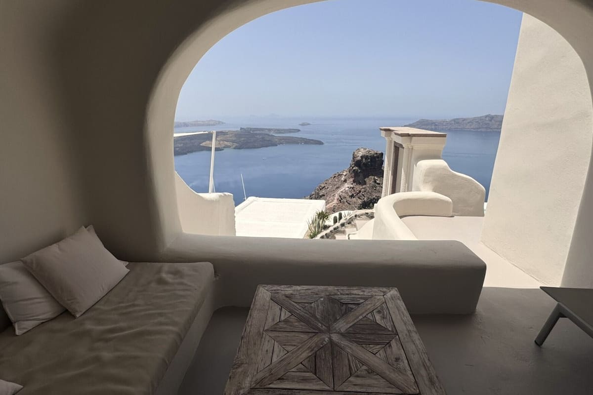 A room with a white sofa and a wooden coffee table, a large arched window offering a view of the sea and rocky landscape.