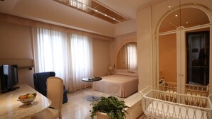 A hotel room with a bed, a desk with a bowl of fruit, a chair, a small table, a potted plant, and a crib. - Hotel Europa (Foggia)