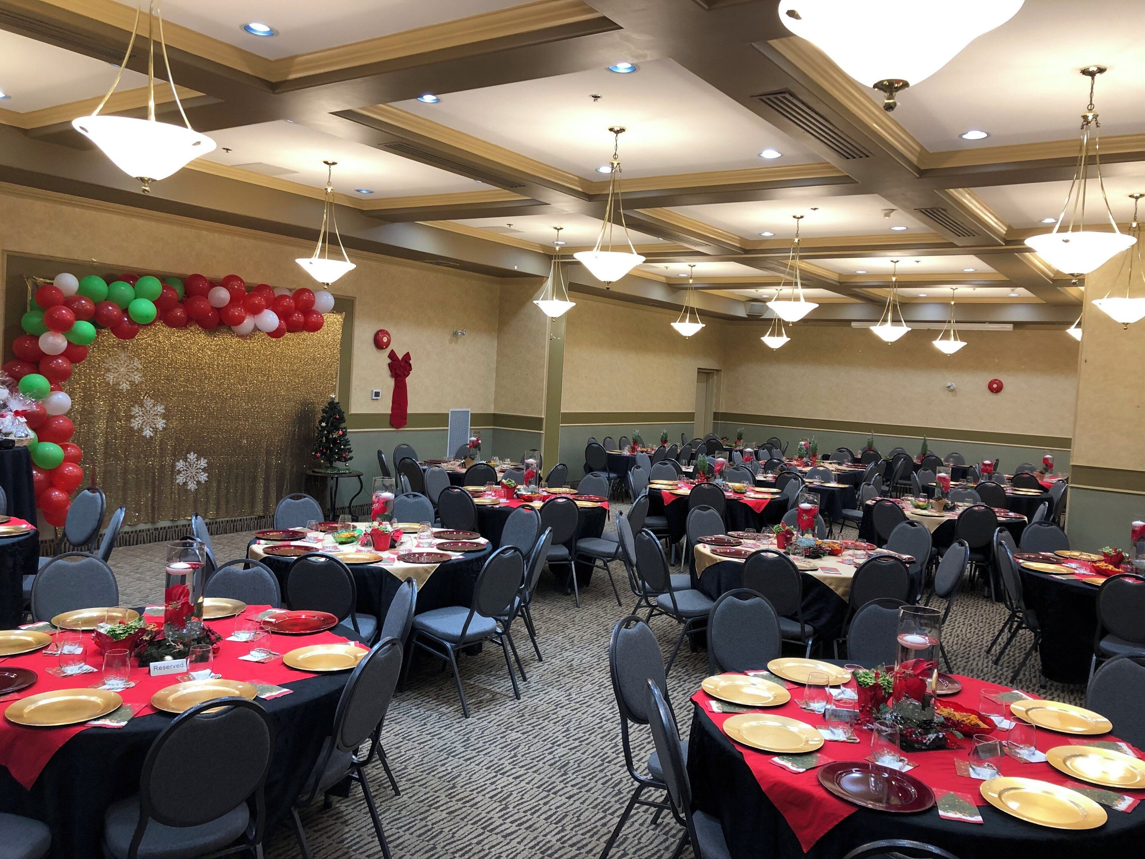 Un salón de banquetes con mesas dispuestas para un evento formal, con decoraciones en rojo y dorado, incluyendo un árbol de Navidad y un arco de globos.