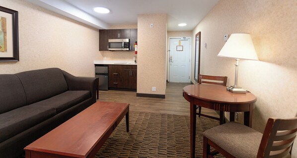 Habitación de hotel con sofá, una mesa de madera, una lámpara, zona de cocina con microondas y una puerta que da a otra habitación.