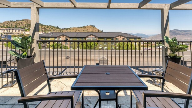 A covered outdoor seating area with a table and benches, overlooking a parking lot and buildings in the background.