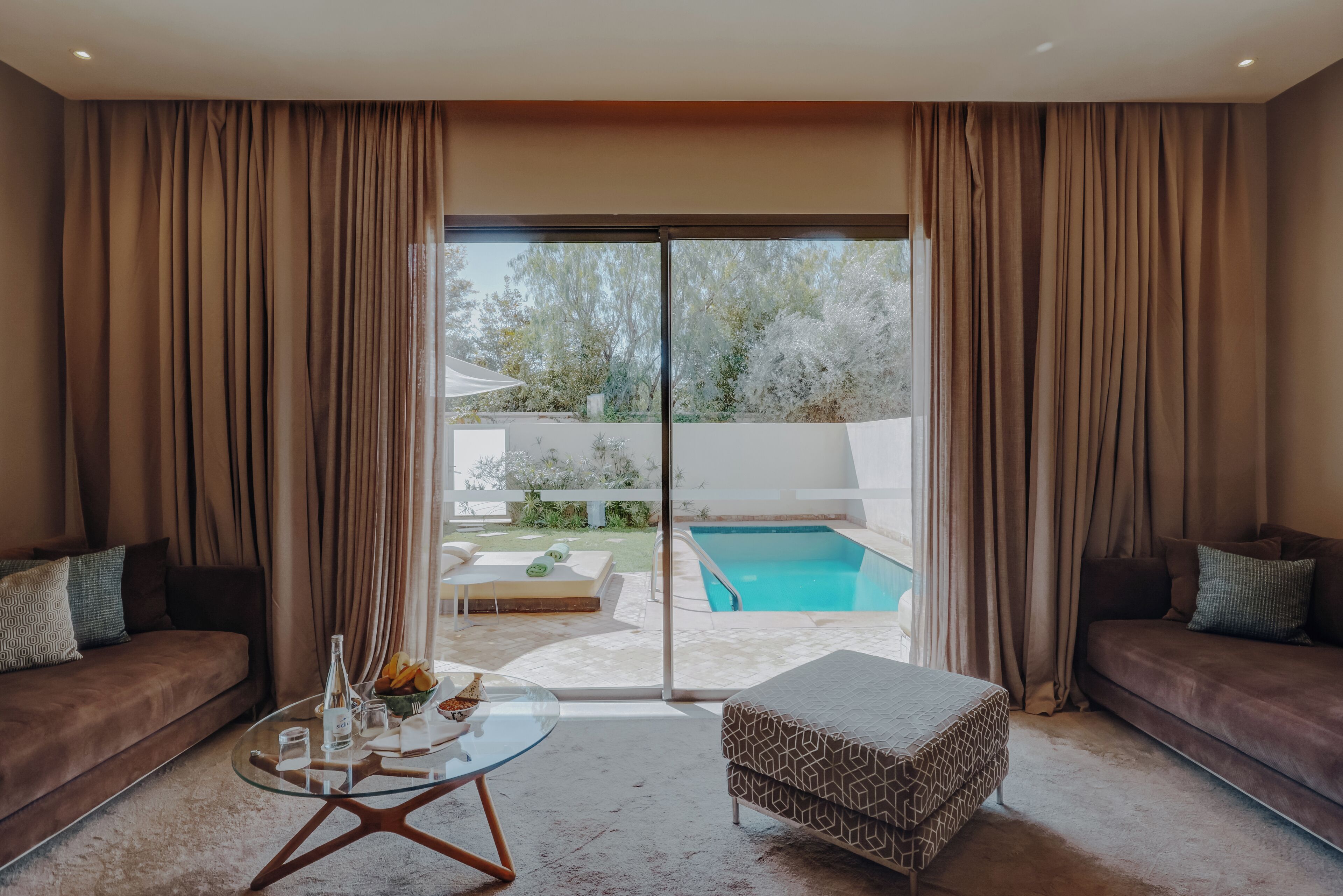 A modern living room with a glass coffee table, a sofa, and an ottoman. There is a sliding glass door leading to a pool area.