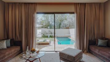 A modern living room with a glass coffee table, a sofa, and an ottoman. There is a sliding glass door leading to a pool area.