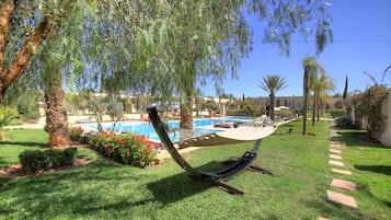 Relax in a serene outdoor setting surrounded by lush greenery and palm trees, featuring a hammock, sun lounger, and swimming pool.