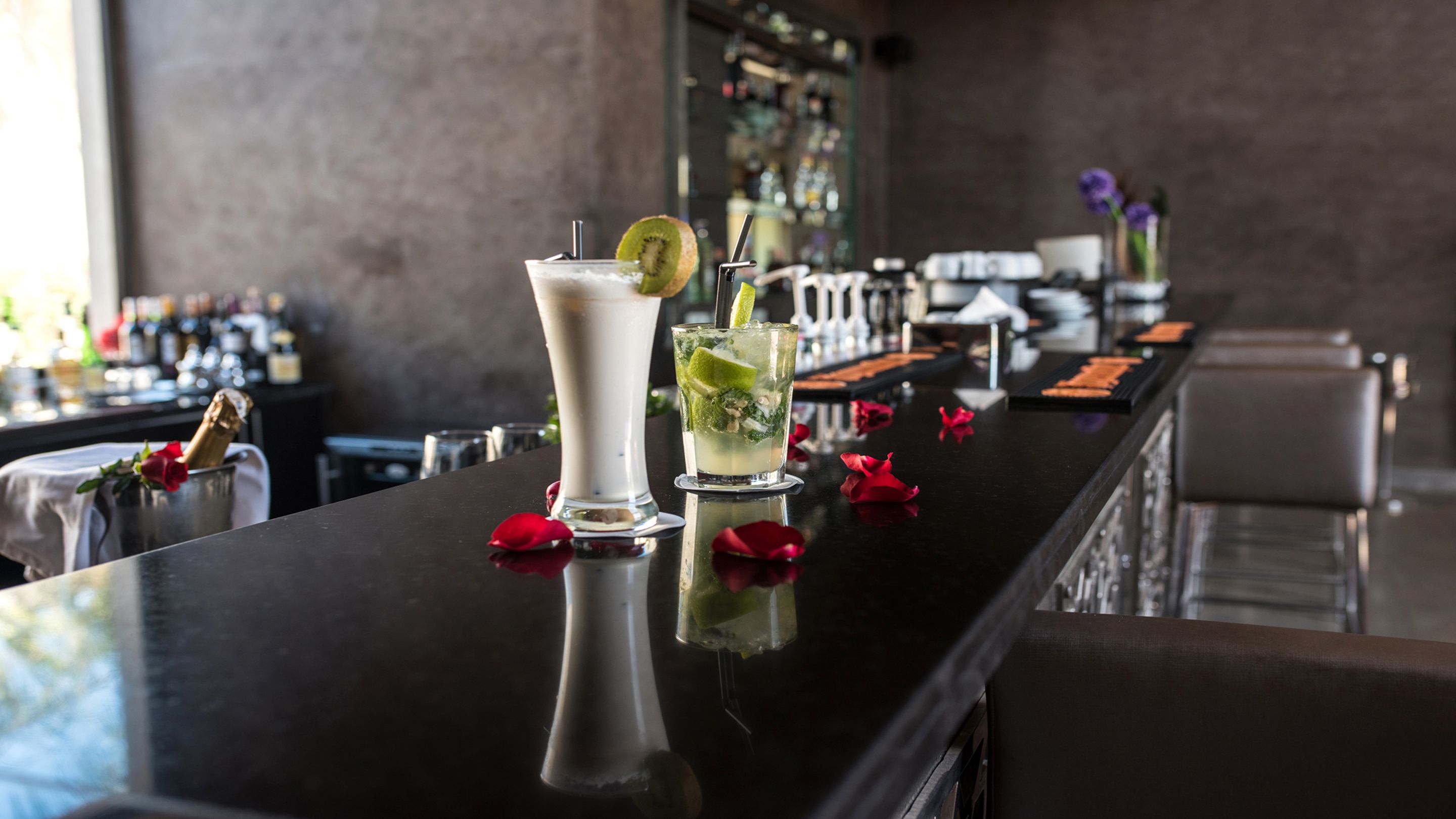 Un espace bar doté d’un comptoir noir poli, de deux cocktails et d’une bouteille de champagne.