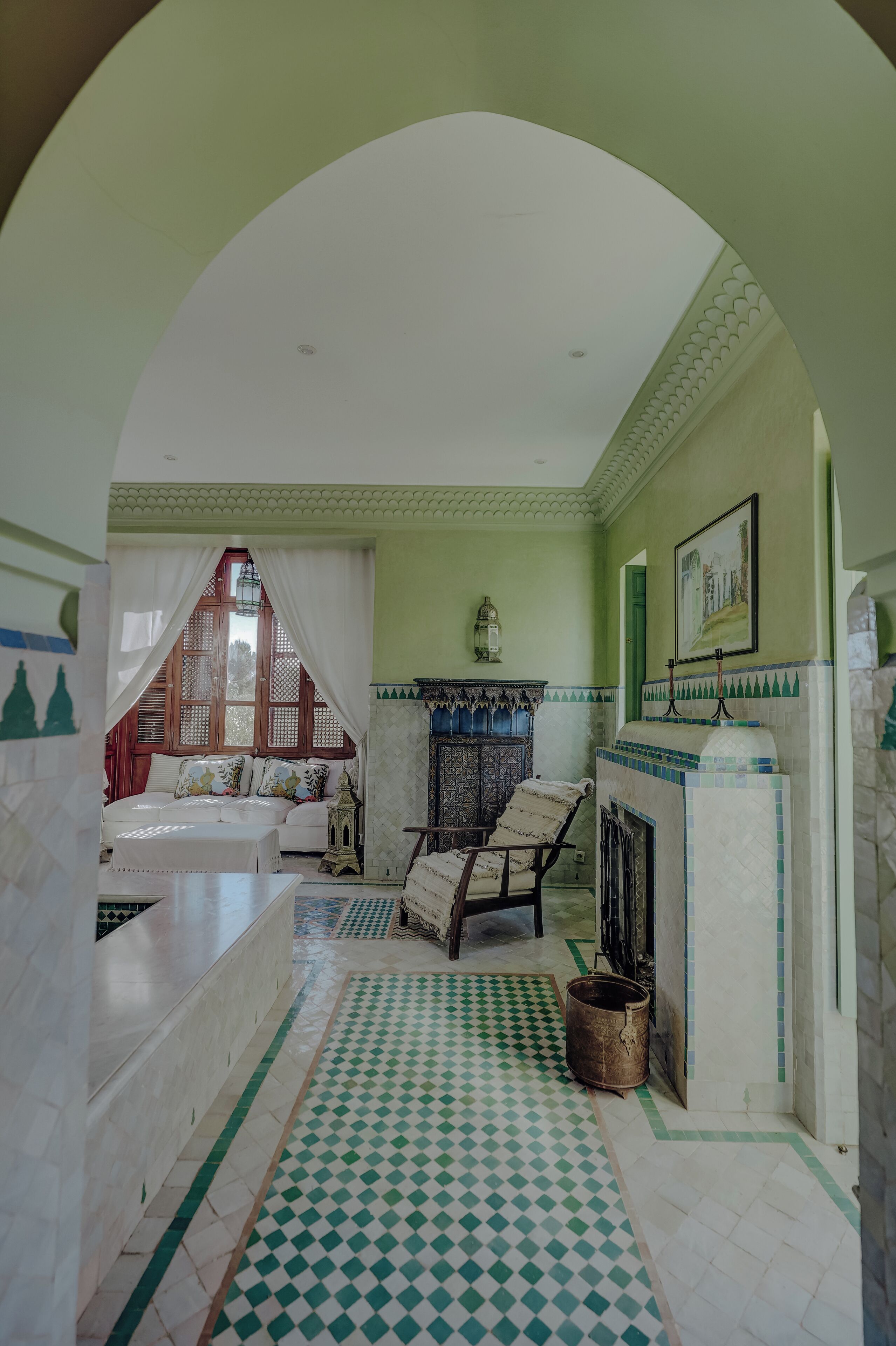 Una habitación con piso a cuadros, una cama con sábanas blancas, una silla, una mesita y un estante fijado a la pared.