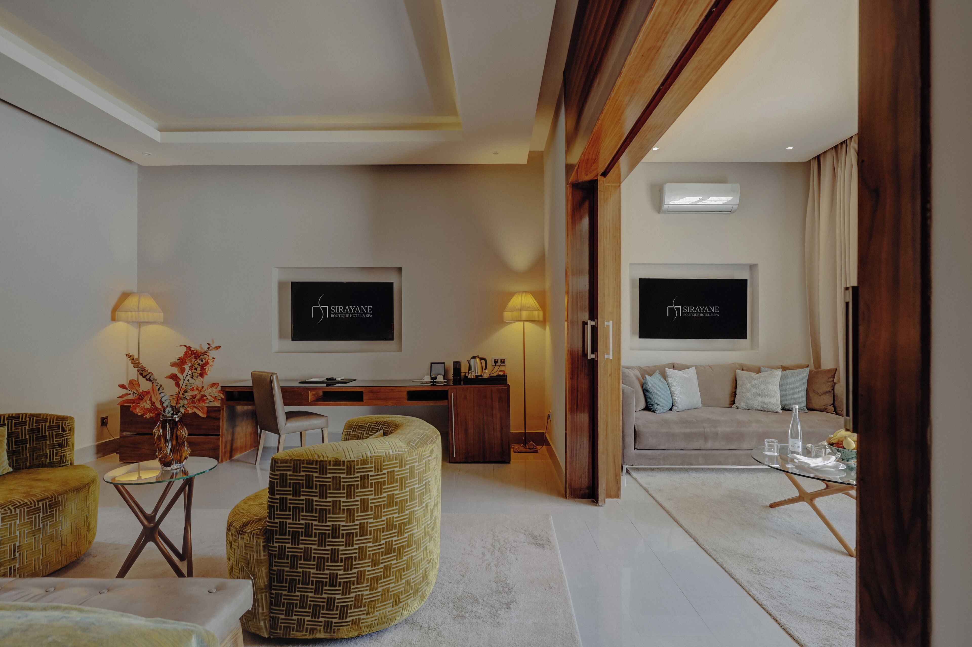 Quarto de hotel moderno com sofá, duas poltronas, mesa de centro de vidro, escrivaninha e duas televisões de tela plana fixadas na parede.