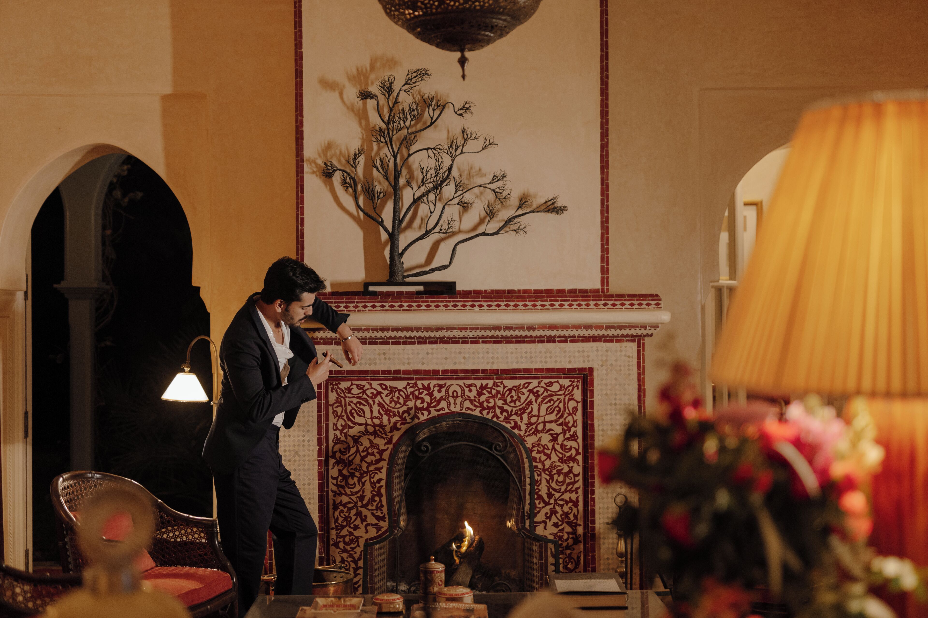 A man in a suit standing by a fireplace with a decorative tree sculpture above it.