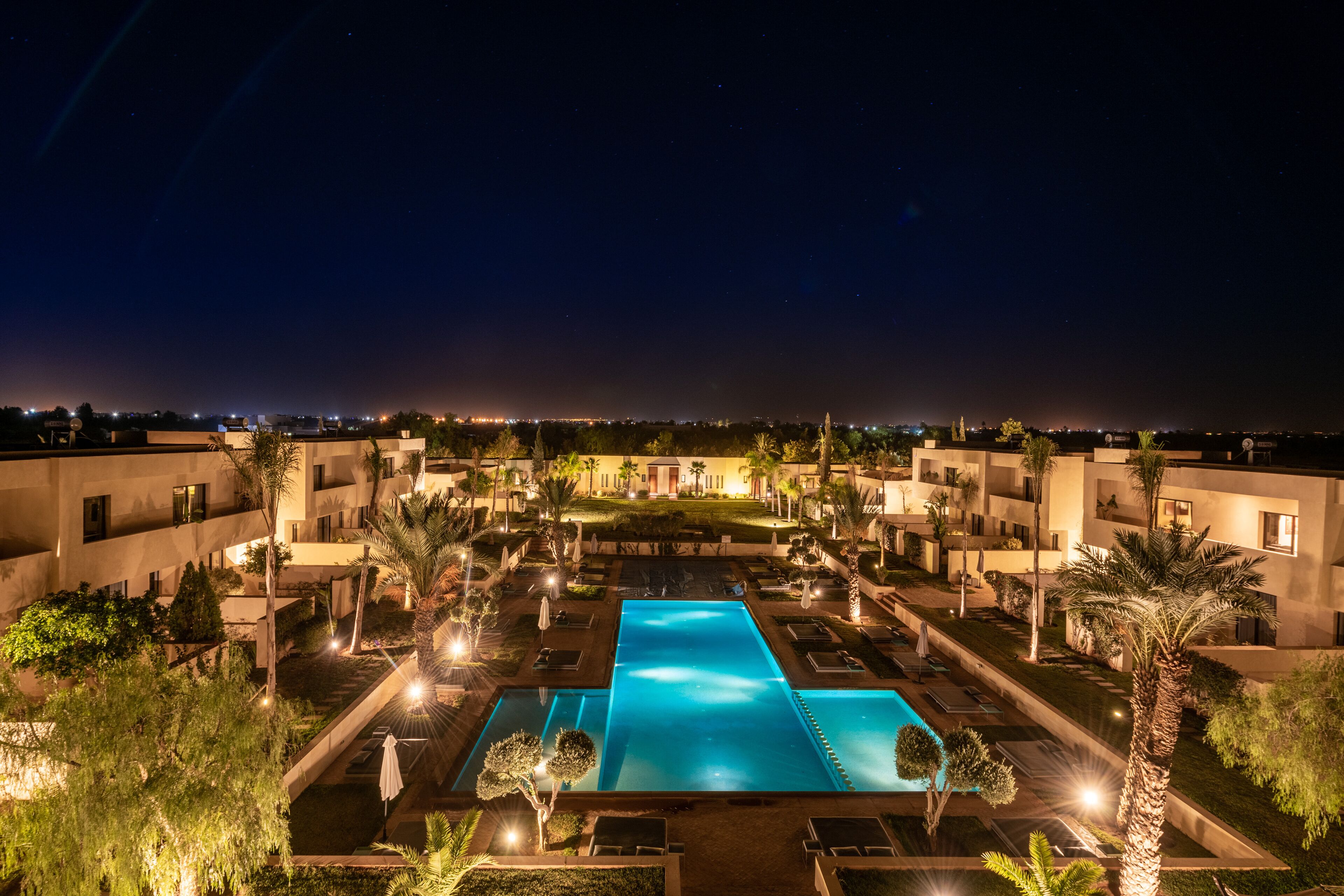 Vista noturna de um resort com piscina central, rodeada por palmeiras e caminhos bem iluminados.