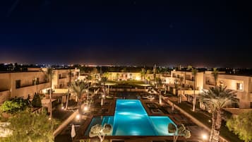 Vista nocturna de un complejo turístico con una piscina central, rodeado de palmeras y senderos bien iluminados.