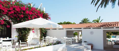 Un espace extérieur aménagé avec des sièges blancs, comprenant des chaises et des tables blanches, entouré d’un mur blanc et surmonté d’un toit recouvert de tuiles rouges.