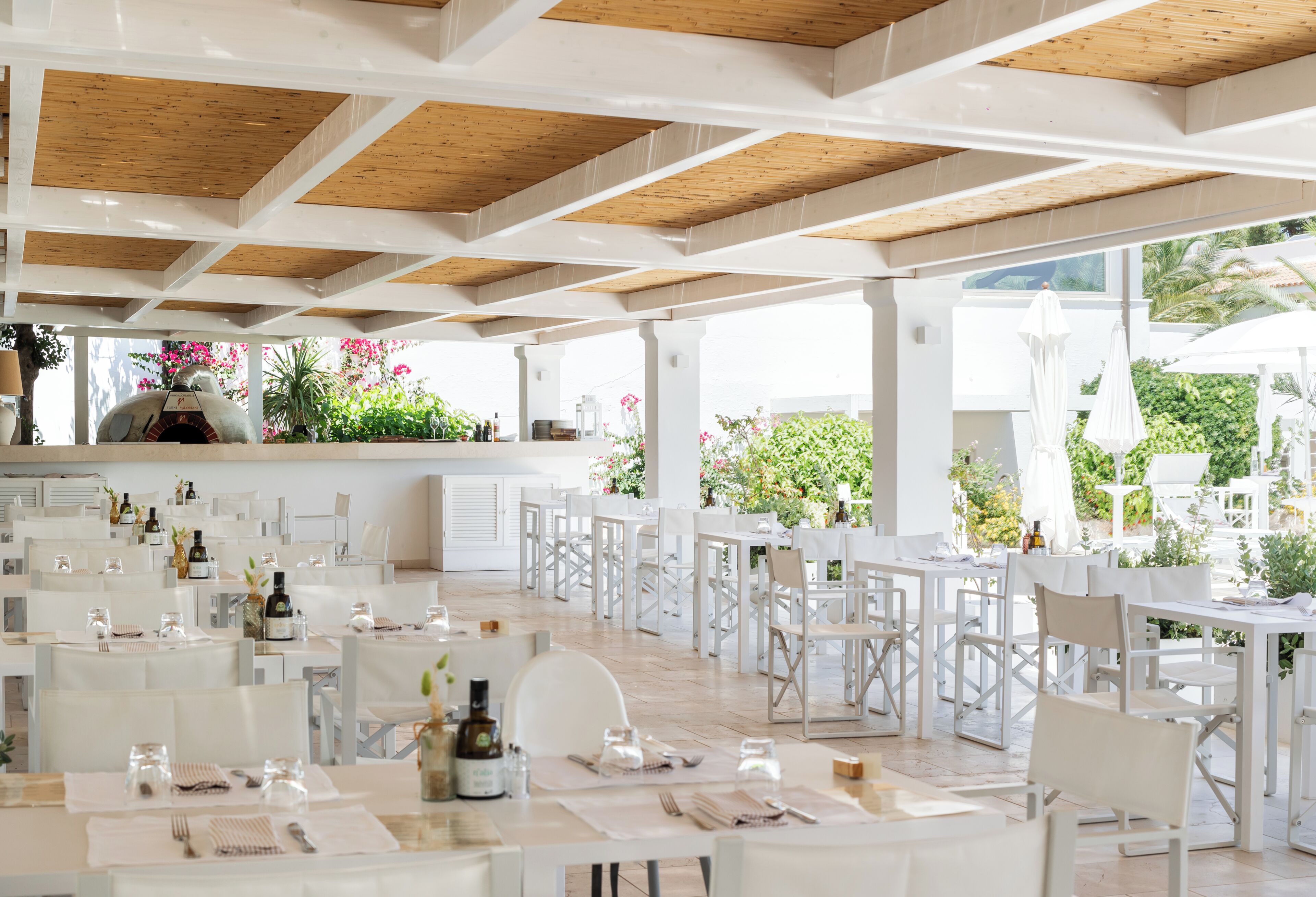 Zona pranzo all'aperto con tavoli e sedie bianche, un tetto in legno e vista su piante e fiori.