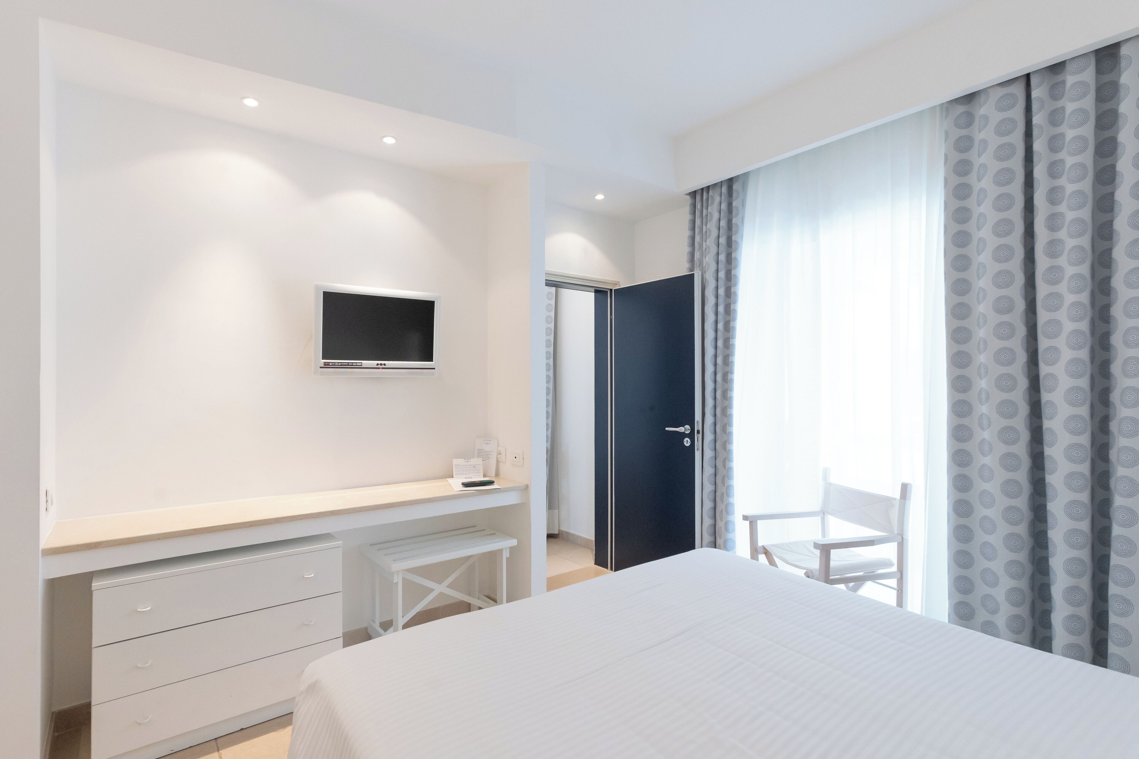A bedroom with a bed, a desk with drawers, a television, and a door leading to a balcony with a chair.