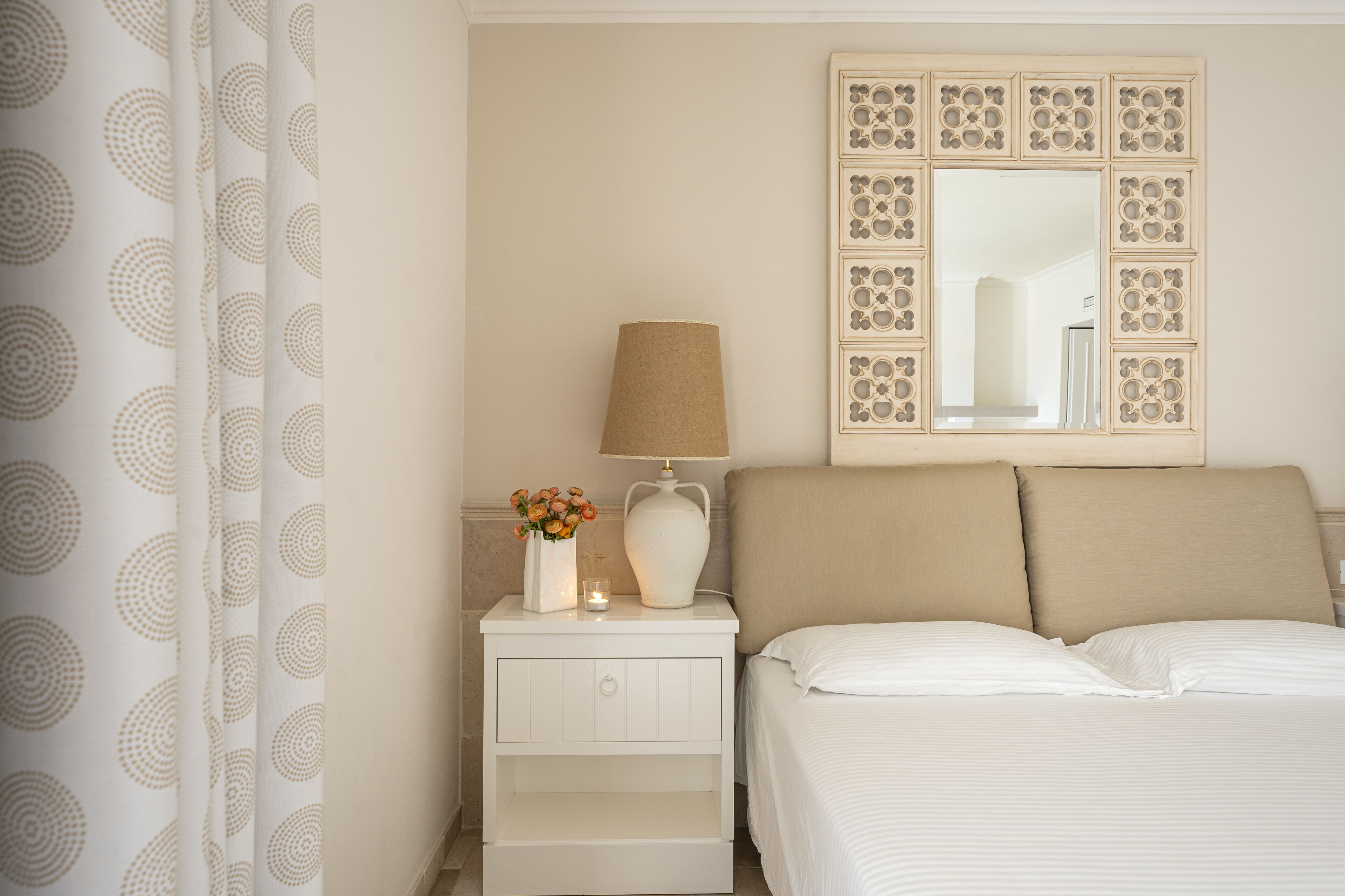 A neatly arranged bedroom with a bed, a nightstand with a lamp, a vase with flowers, and a decorative mirror on the wall.