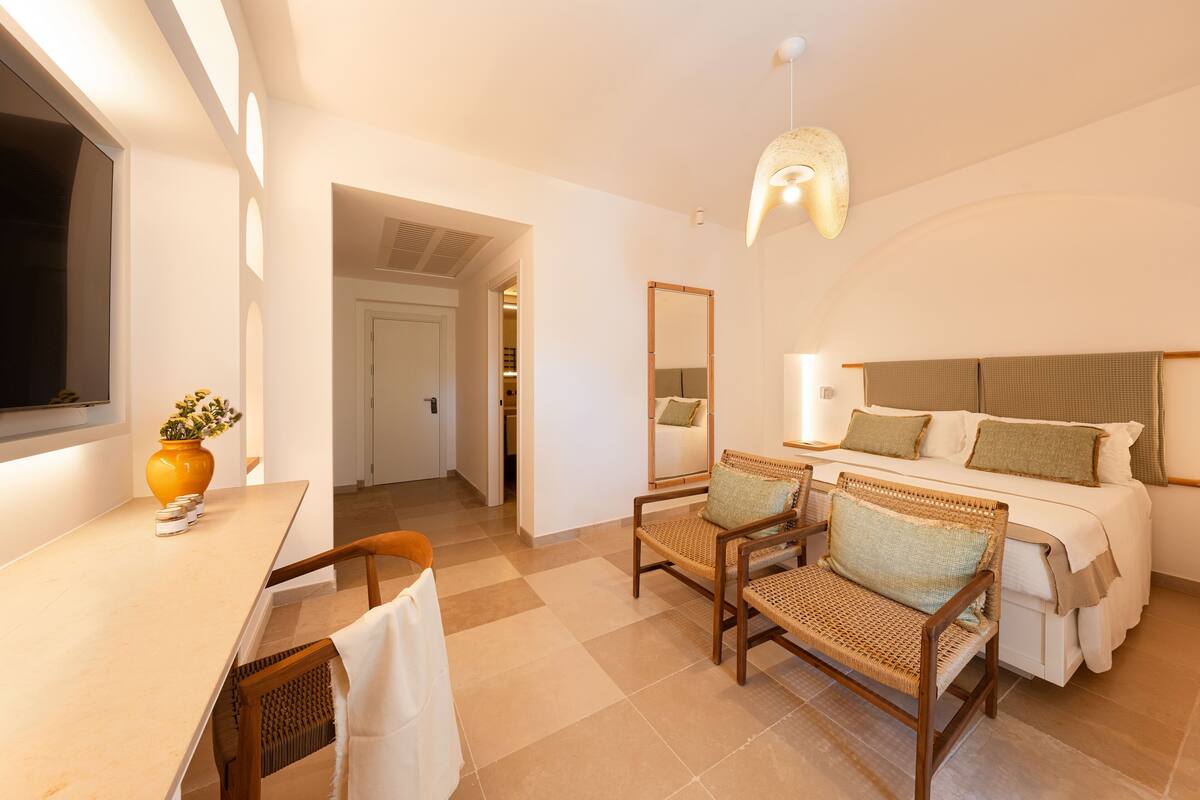 A hotel room with a bed, two wicker chairs, a desk with a vase, a mirror, and a ceiling light fixture.