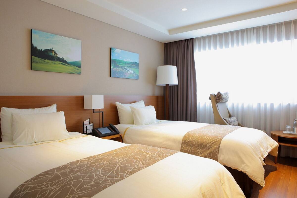 Una habitación de hotel con dos camas, una mesita de noche con lámpara, una silla y una obra de arte en la pared.