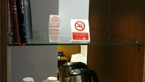 Habitación de hotel con repisa de vidrio que muestra libros, un cartel de prohibido fumar y una estación de café con tazas y hervidor.