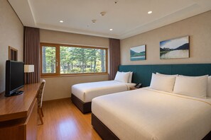 Habitación de hotel con dos camas, un escritorio con televisor y ventana con vistas a árboles.