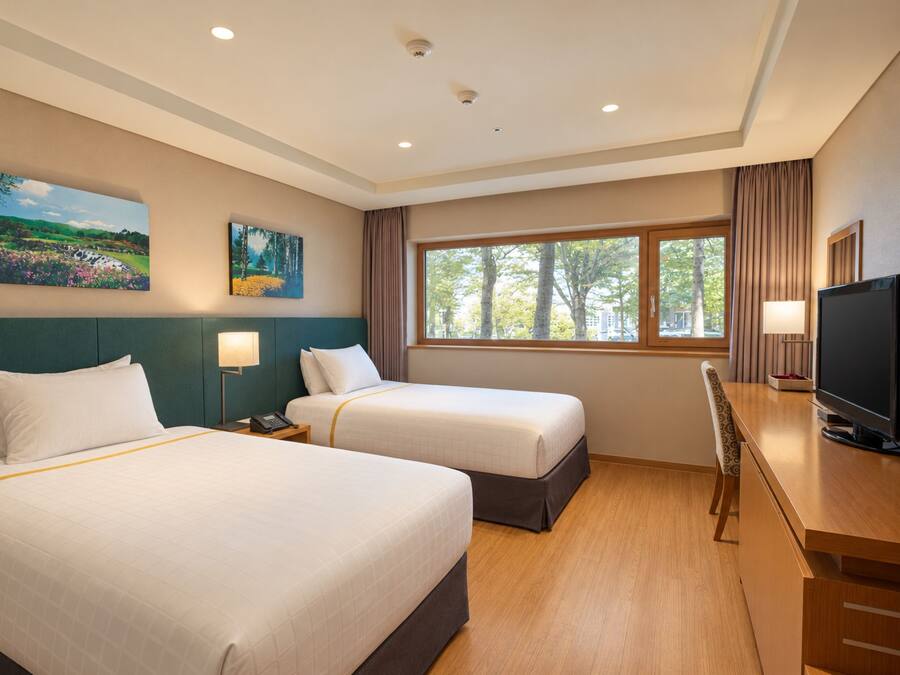 A hotel room with two beds, a desk with a television, and a window with a view of trees.