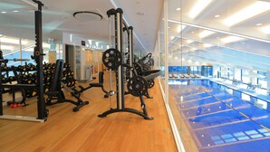 Une salle de sport équipée de matériel de musculation, une piscine bleue et une vue sur la ville grâce à de grandes fenêtres.