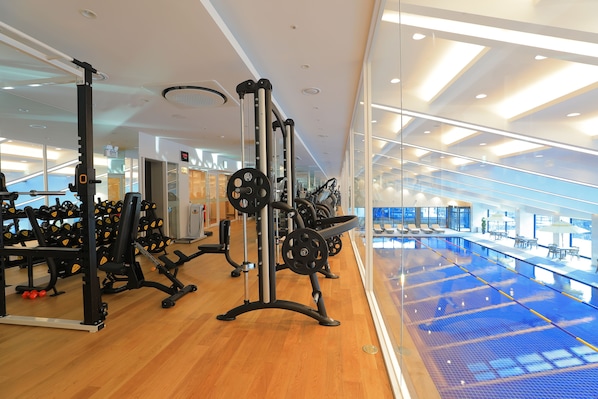 Un gimnasio con equipo de levantamiento de pesas, una piscina azul y vistas de la ciudad a través de grandes ventanales.