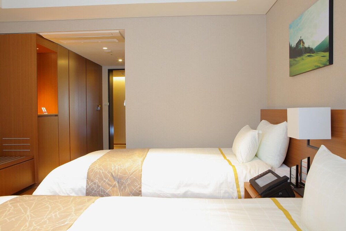 Une chambre d’hôtel avec deux lits, une armoire en bois, un tableau au mur et une table de chevet avec une lampe et un téléphone.