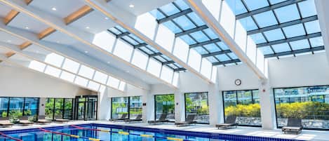 Una piscina interior moderna con techos de vidrio transparente, zonas iluminadas por el sol y sillas de descanso.