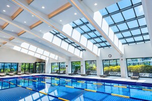 Una piscina interior moderna con techos de vidrio transparente, zonas iluminadas por el sol y sillas de descanso.