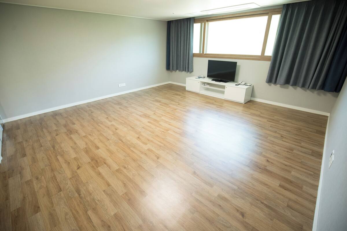 Un salón amplio con suelo de madera, un mueble de entretenimiento blanco y un ventanal con cortinas.