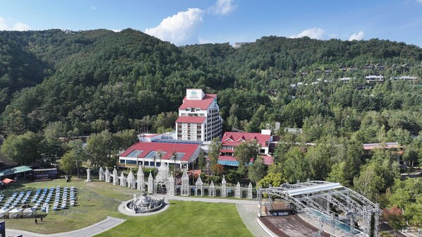 A hotel with a red roof, surrounded by a park with a fountain and white structures.