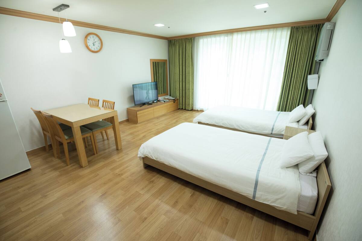 Habitación de hotel con dos camas, un escritorio de madera con sillas, un televisor y un reloj en la pared.