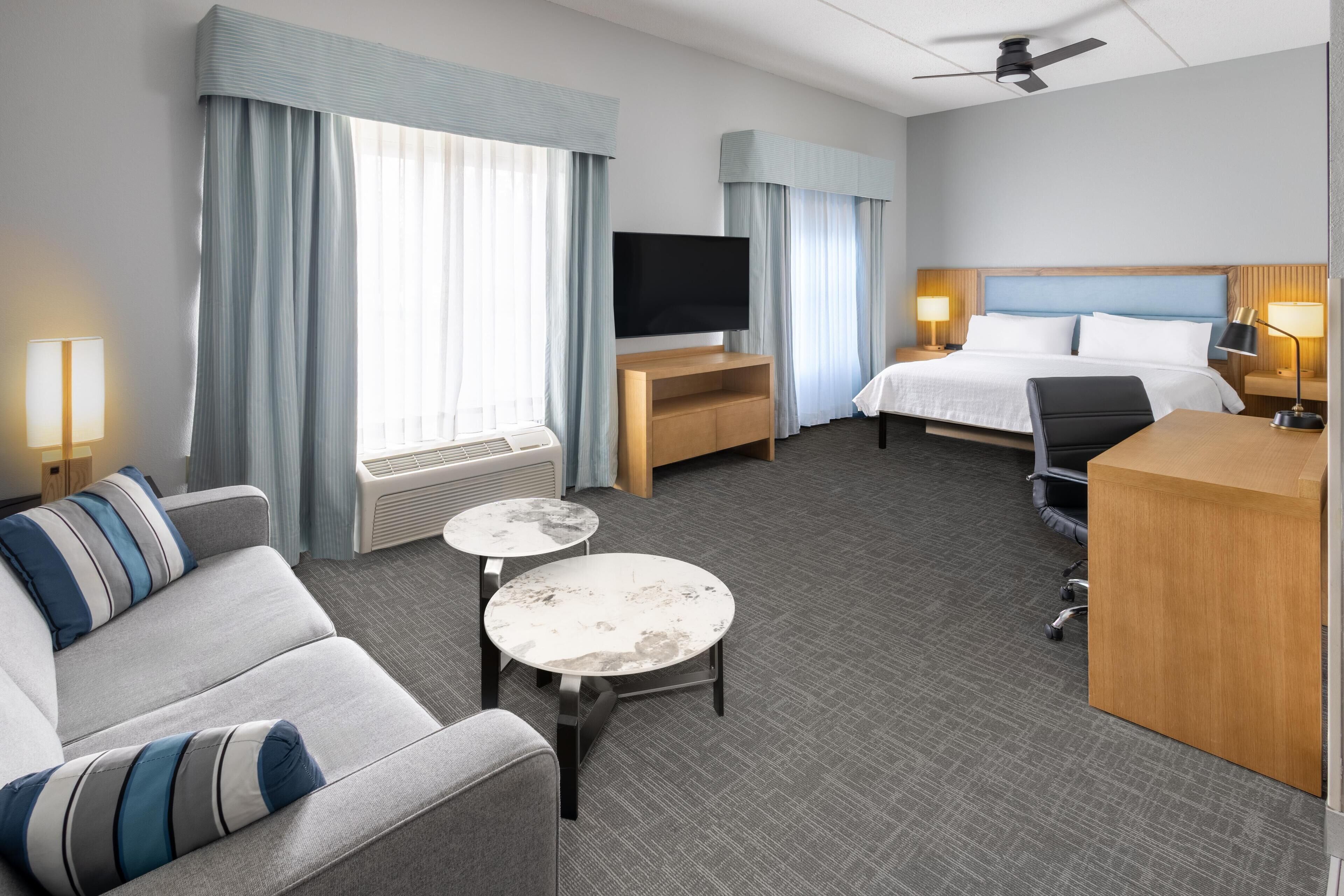 A hotel room with a bed, a desk, a television, a sofa, two small tables, and a ceiling fan.